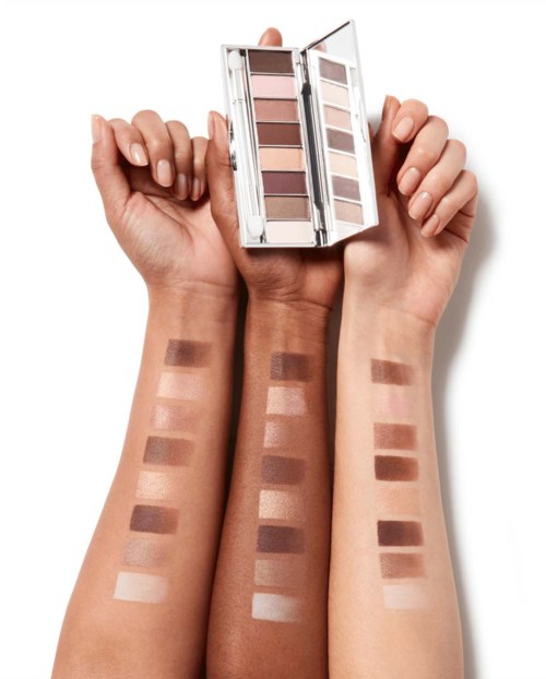 Iemand houdt een oogschaduwpalet met bruin- en beige tinten vast, met swatches op de onderarmen.