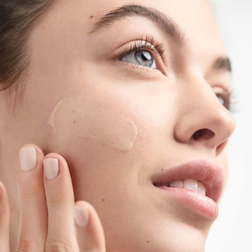 Een vrouw brengt vochtinbrengende crème of foundation aan op haar wang, met een heldere huid en blauwe ogen. Geen make-up zichtbaar.