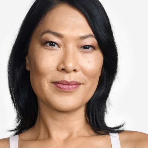 Eine Frau mit schwarzen Haaren und leichtem Lächeln, trägt ein weißes Top, vor einem weißen Hintergrund.