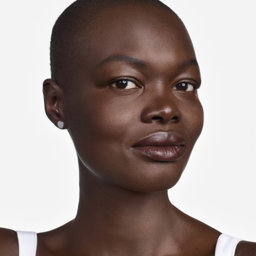 A woman with dark skin and a shaved head looks confidently at the camera. She wears a white top with small earrings.