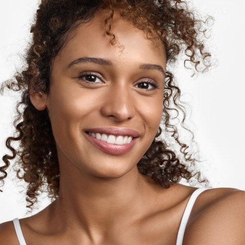 Eine Frau mit lockigem Haar, die lächelt und in die Kamera schaut, trägt ein weißes Top.