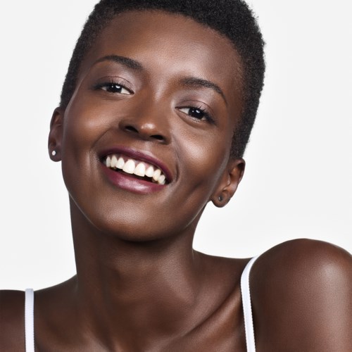 A smiling woman with short curly hair and earrings, wearing a white strap top, against a plain background.