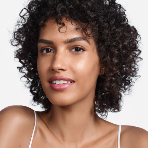 Eine Frau mit lockigem Haar und glatter Haut lächelt sanft, trägt ein weißes Spagettiträger-Top.