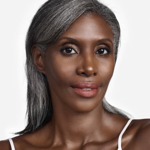 A woman with gray hair and natural makeup, wearing a white strap top, smiling slightly. Simple background.