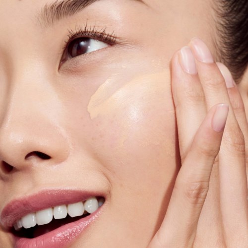 Een vrouw brengt foundation of concealer aan op haar wang, glimlacht zachtjes, toont een heldere huid en natuurlijke schoonheid.