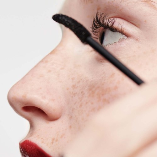 Een persoon brengt mascara aan op de wimpers met een applicator, gericht op oog make-up. Ze hebben sproeten en rode lipstick.