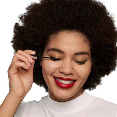 Een vrouw met krullend haar glimlacht en brengt eyeliner aan. Ze draagt rode lipstick en een witte shirt.