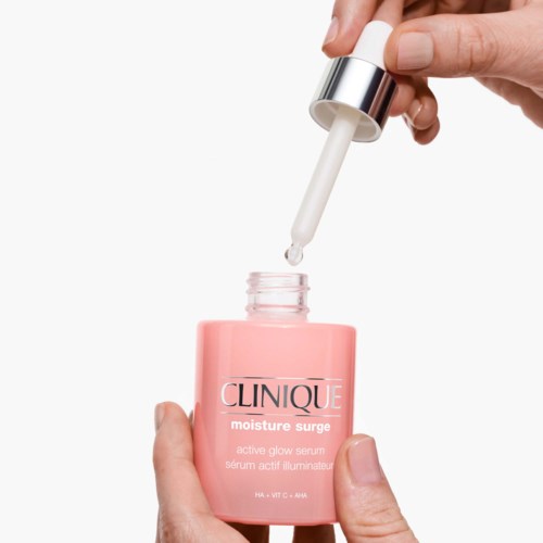 Een hand houdt een roze fles met 'Clinique moisture surge' en een pipet die serum afgeeft.