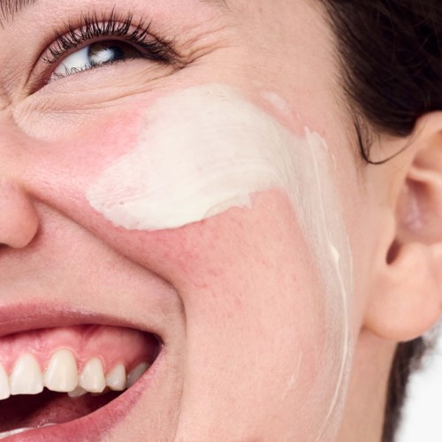 Een vrouw brengt crème aan op haar wang, glimlacht met zichtbare tanden en deel van haar oog en gezicht.