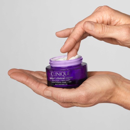 Een persoon schept crème uit een paarse pot met 'Clinique' erop met een hand. De achtergrond is eenvoudig en grijs.