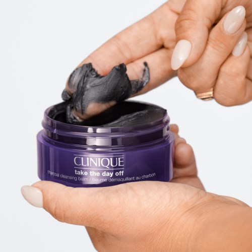 Een hand haalt een zwarte reinigingsbalsem uit een paarse pot met het label 'Clinique Take the Day Off.'