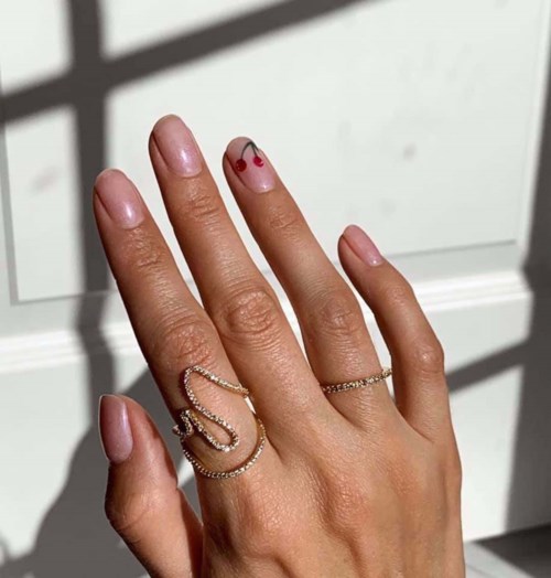 Eine Hand mit Ringen, Nagelkunst und einer kleinen Kirschtätowierung am Mittelfinger.