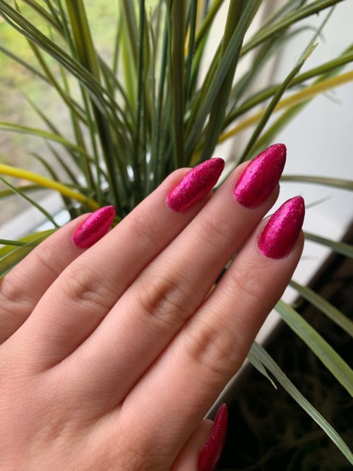Vijf vingers met glanzende donkerroze nagellak dichtbij groene planten.