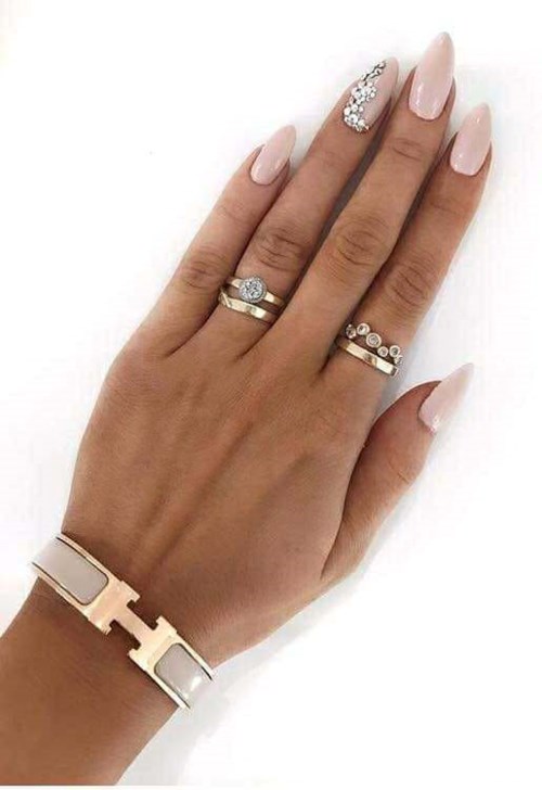 Hand mit hellrosa Nägeln, Ringen und weißem Armband auf weißem Hintergrund.