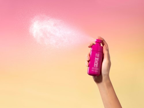 En hånd sprayer en rosa aerosolkanna med hvitt pulver mot en gradient rosa og gul bakgrunn.
