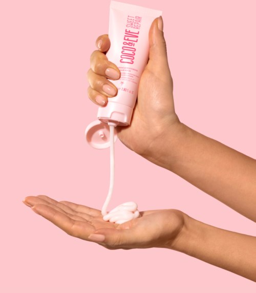 A hand holds a pink tube with cream, pouring onto an open palm, against a pink background.