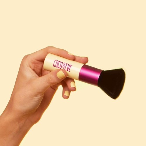 Een hand houdt een make-up penseel met een roze en zwart handvat, en de tekst 'COCO & EVE' op het handvat.