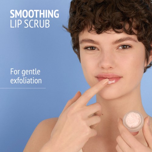 A young woman with short curly hair applies lip scrub, smiling softly, against a light blue background.