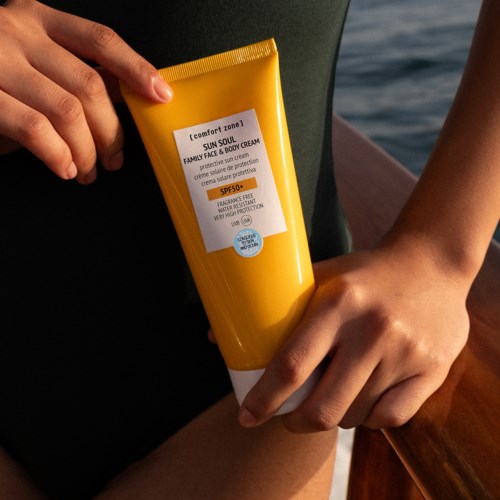 Person holding a yellow tube of sun protection face and body cream near water. The cream has SPF 50.