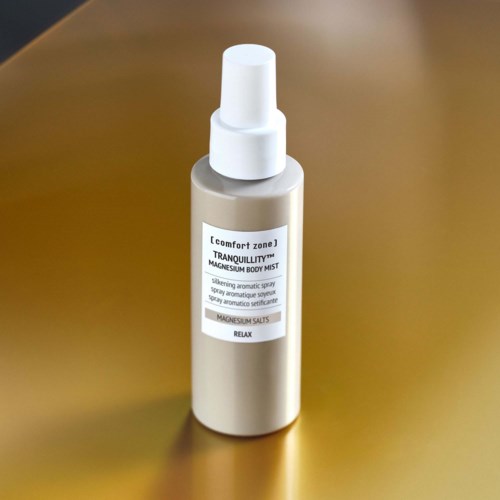 En silver sprayflaska märkt magnesium kroppsspray för avkoppling, med vit kork, på en guldig yta.