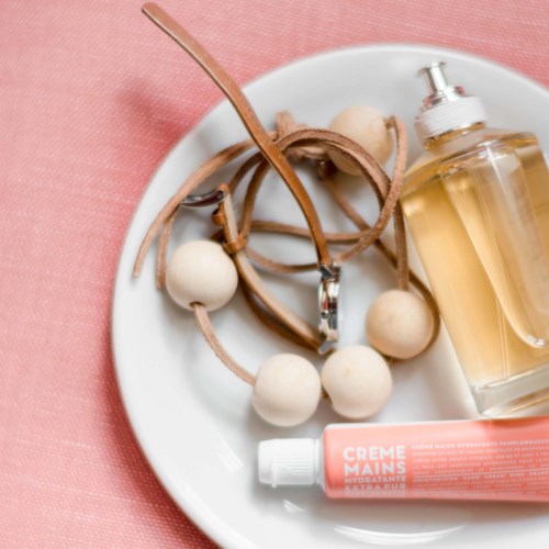 A white plate holds jewelry, a perfume bottle, and hand cream, all on a pink background.