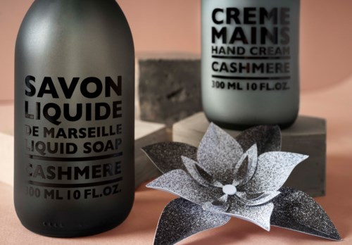 Two dark bottles of liquid soap and hand cream with black and silver decorative flowers on a pink surface.