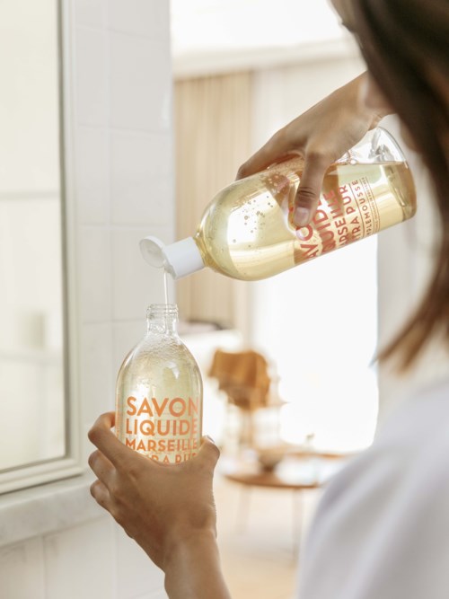 Someone is pouring liquid soap into a small bottle. The room is bright with a cozy atmosphere.
