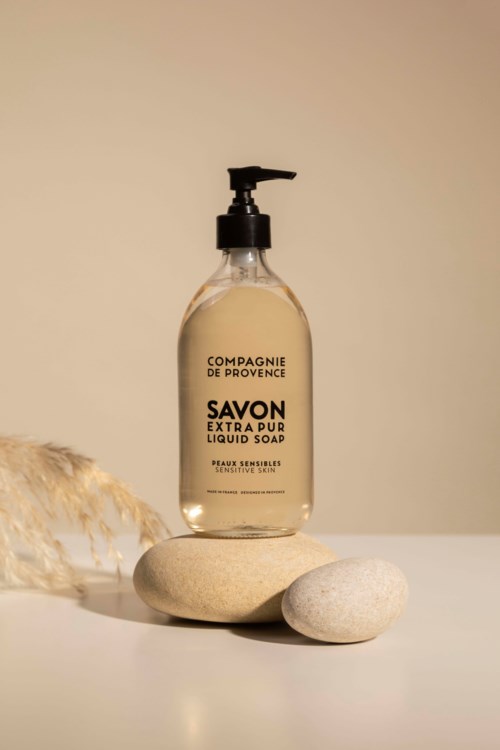 A clear liquid soap bottle sits on stones, with a soft-colored background and a plant in the corner.