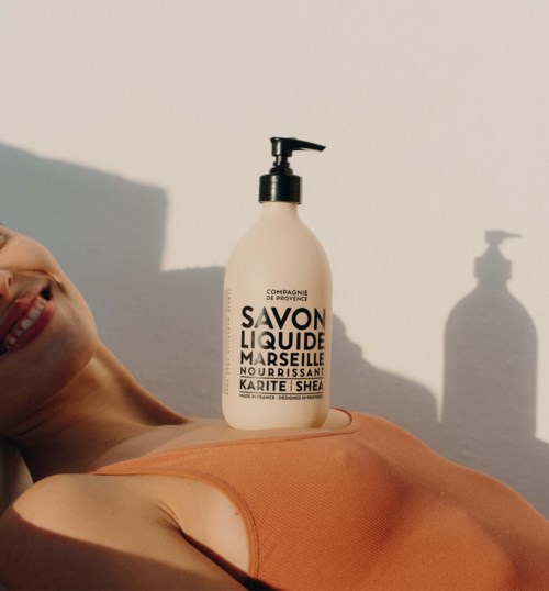 A smiling woman lying down with a bottle of liquid soap on her chest, shadows visible on the wall.