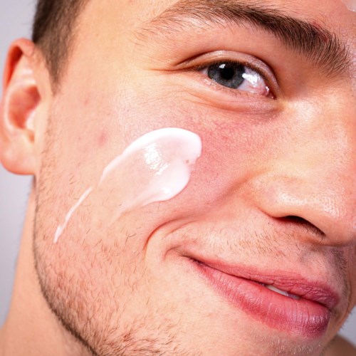 A man applies white cream or lotion on his cheek, smiling gently, close-up of his face.
