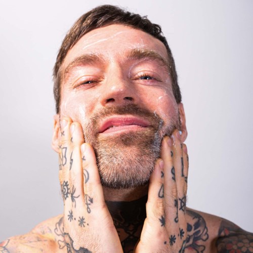 A tattooed man with a beard, smiling softly, touching his face with both hands.