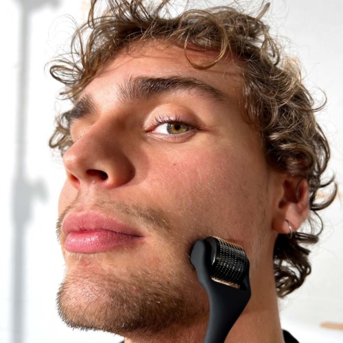 A young man with curly hair and light skin uses a facial roller on his jawline.