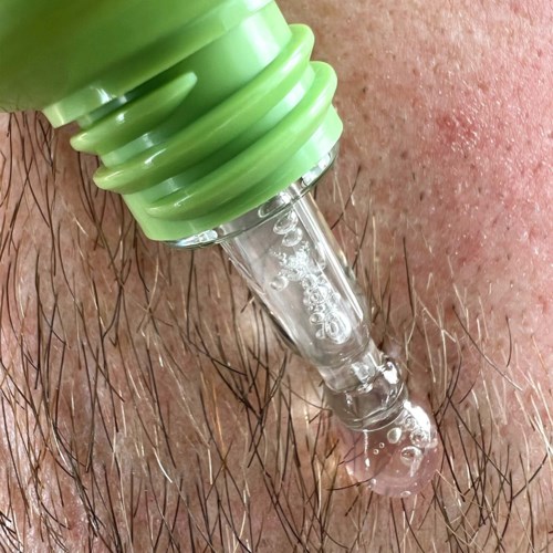 A green dropper administers a liquid onto skin with hair, creating bubbles and a small droplet at the end.
