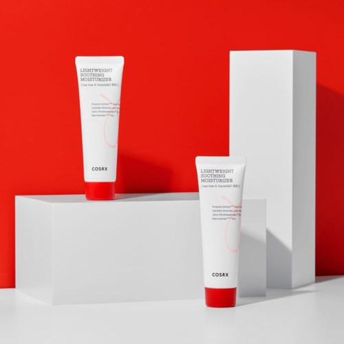 Twee witte tubes hydraterende crème met rode deksels op witte blokken voor een rode achtergrond.