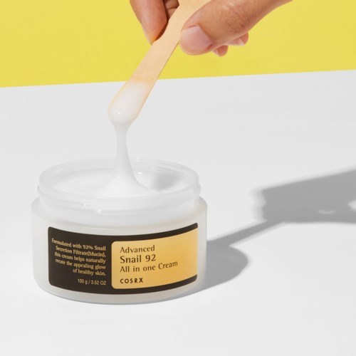 A hand with a stick dips into a jar of white cream labeled 'Snail 92 All in one Cream.' The background is yellow.