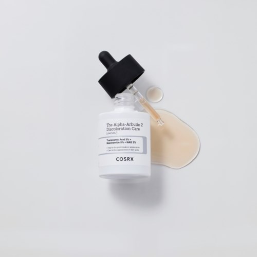 Een kleine huidverzorgingsfles met pipet en een kleine witte dop, met een druppeltje beige vloeistof ernaast.
