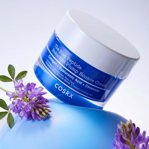 En blå boks av COSRX Blue Peptide Bounce Cream på en blå overflate med lilla blomster i nærheten.