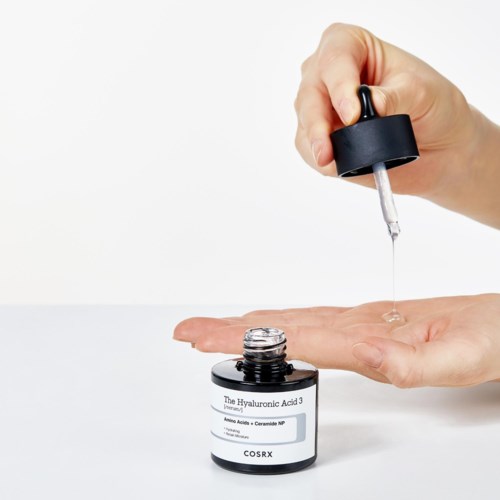 Een hand met een pipet druppelt heldere serum op een andere hand, met een klein potje serum in de buurt.