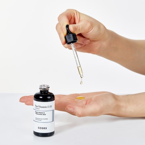 Een hand gebruikt een pipet om vitamine C in de palm van de andere hand te druppelen. Er staat een kleine fles in de buurt.