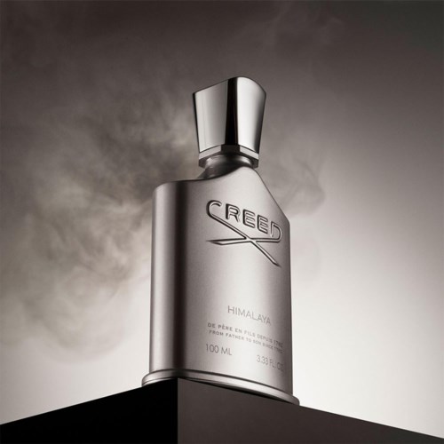 En elegant silverflaska märkt 'Creed Himalaya' står på en svart platta med en molnig, mörk himmel i bakgrunden.