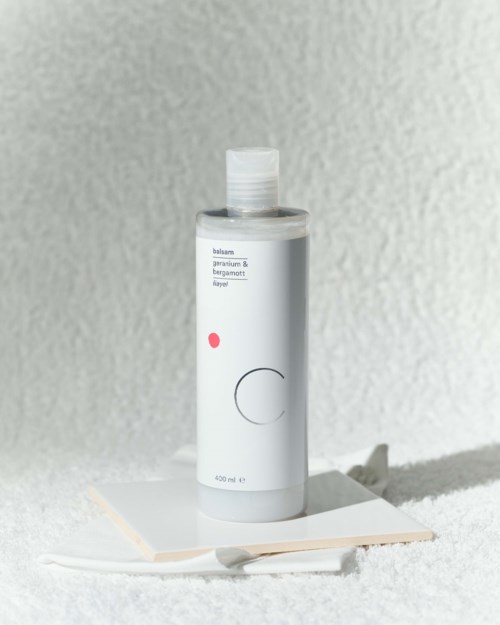 En vit flaska med enkel design står på en vit yta, mot en strukturerad grå vägg.