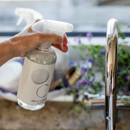En hand sprayar rengöringsmedel från en genomskinlig flaska på en blank metallkran.