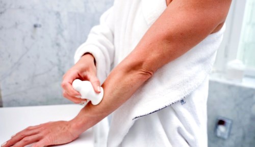 Eine Person trägt Creme oder Lotion auf den Arm auf, während sie ein weißes Handtuch trägt, in einem hellen Badezimmer.