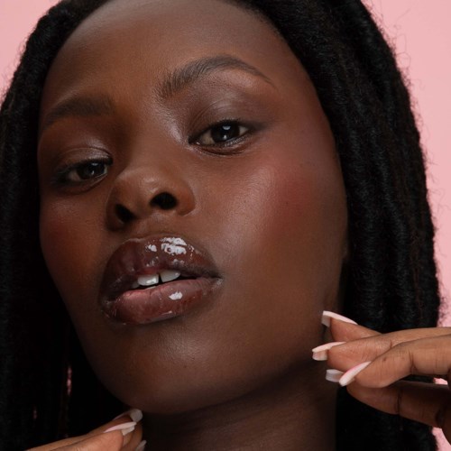Close-up van een donkerhuidige vrouw met glanzende lippen en dreadlocks, die haar gezicht aanraakt met roze achtergrond.