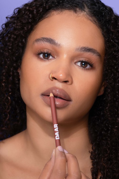 Een vrouw met kroeshaar brengt nude lippenstift aan, houdt het met haar lippen en kijkt naar de camera.