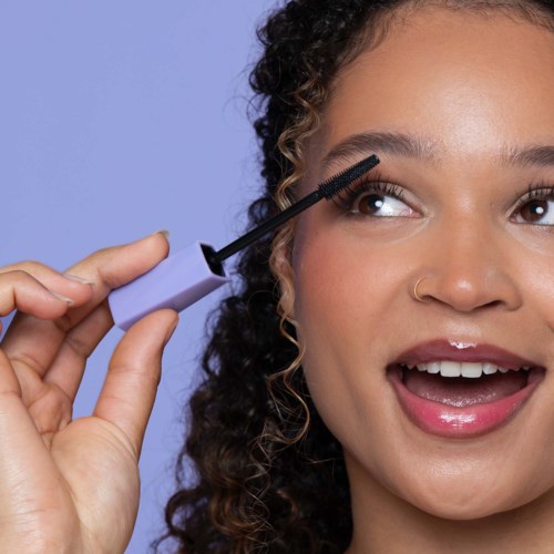 Een vrouw kijkt naar haar wenkbrauwborstel met haar mond open, houdt een mascara borsteltje bij haar oog.