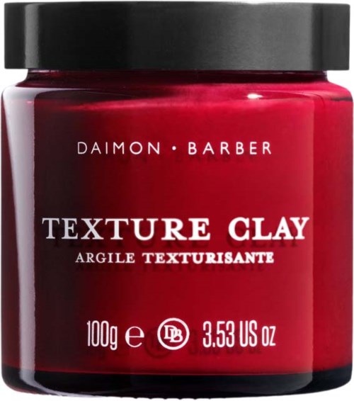 En röd burk med svart lock märkt 'Daimon Barber Texture Clay' för hårstyling.