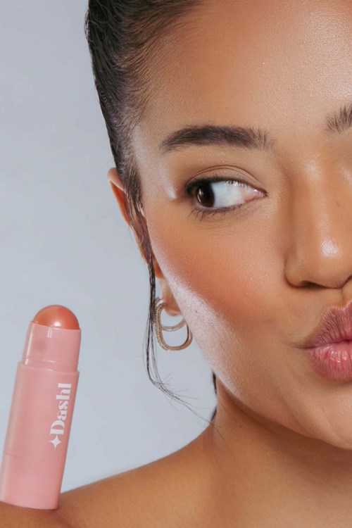 Een vrouw houdt een roze deodorant vast, maakt een speels gezicht en pout haar lippen, kijkt zijwaarts.