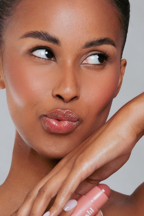 A woman with shiny skin, thinking or puzzled, holding a tube, possibly lipstick, with a neutral background.
