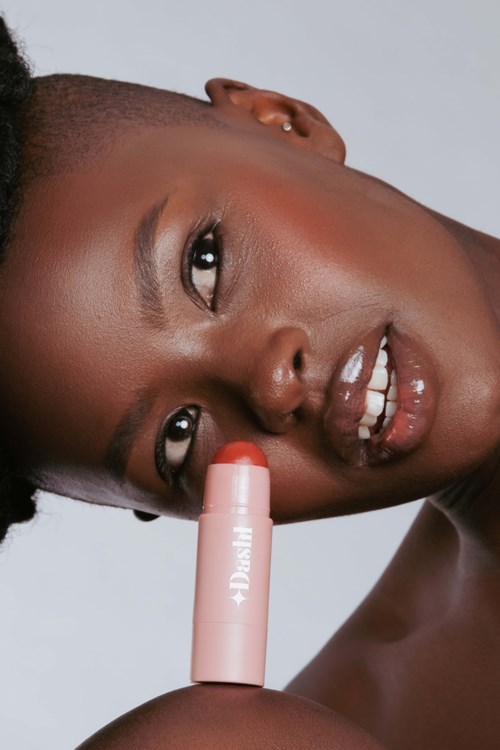 Een vrouw met een donkere huid houdt lippenstift dicht bij haar gezicht. Ze glimlacht breed en kijkt naar de camera.